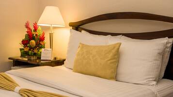 A neatly made bed with white and beige linens, a wooden headboard, and a nightstand with a lamp and a floral arrangement.