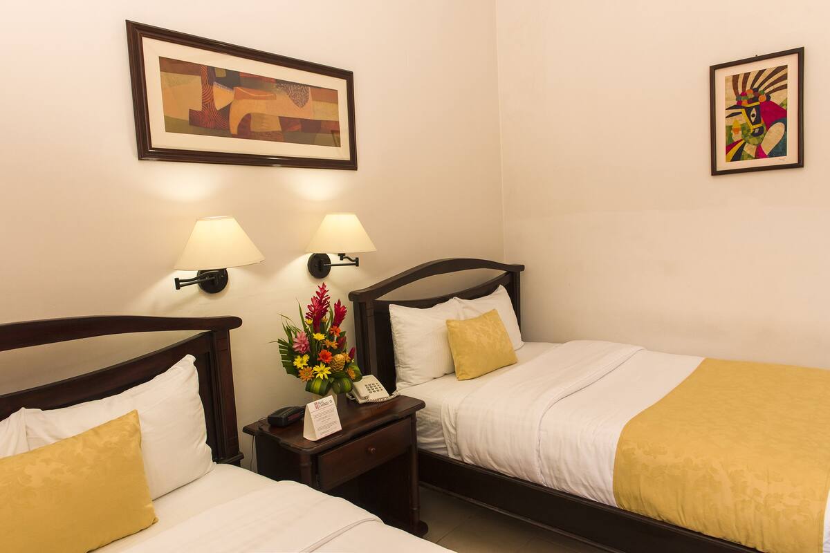 Quarto de hotel com duas camas, uma mesa de cabeceira com abajur, telefone e um quadro na parede.