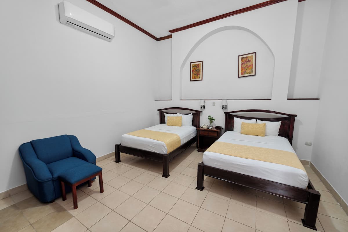 Quarto de hotel com duas camas de solteiro, uma poltrona azul e um aparelho de ar condicionado.
