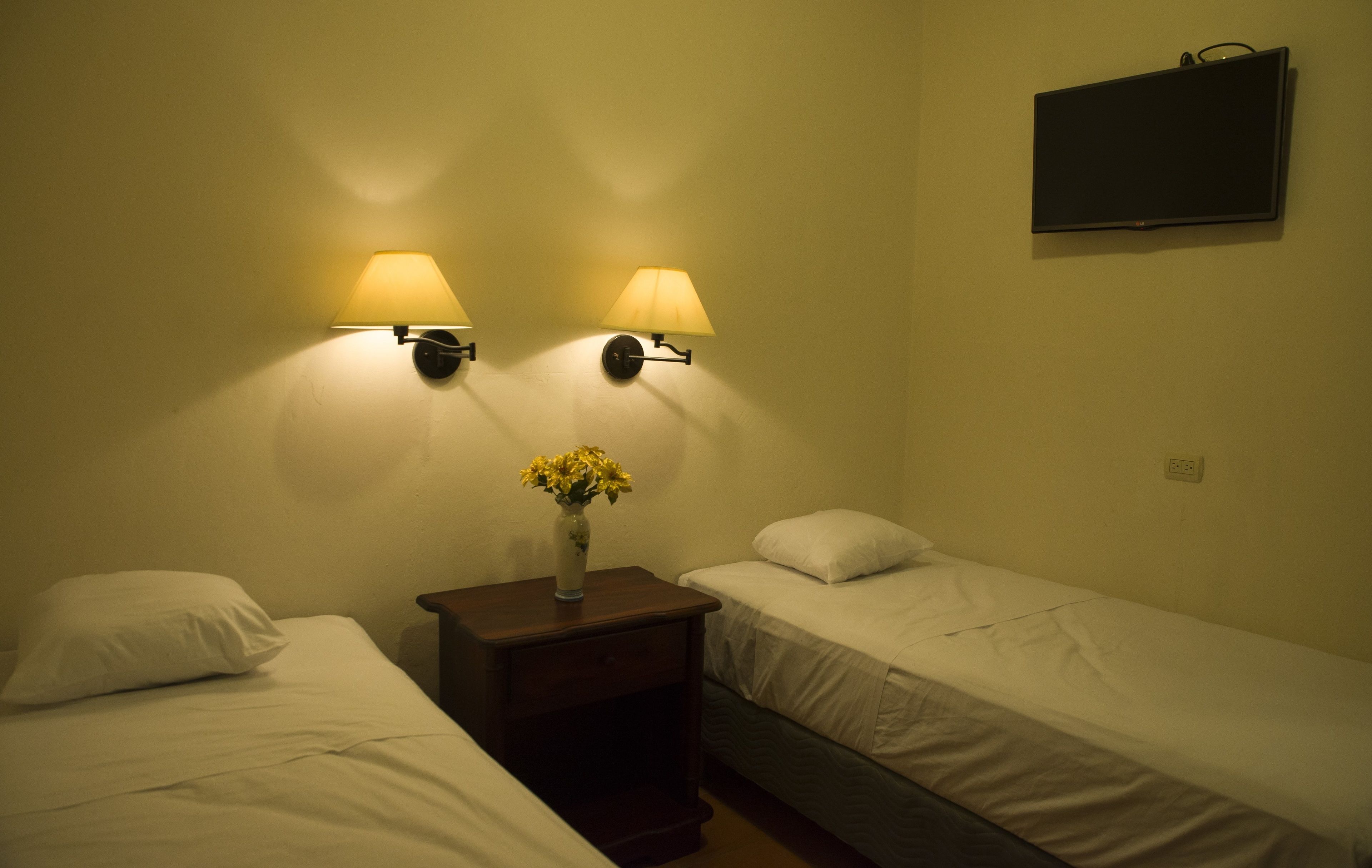A small, dimly lit room with two single beds, a nightstand with a vase of flowers, and two wall-mounted lamps.