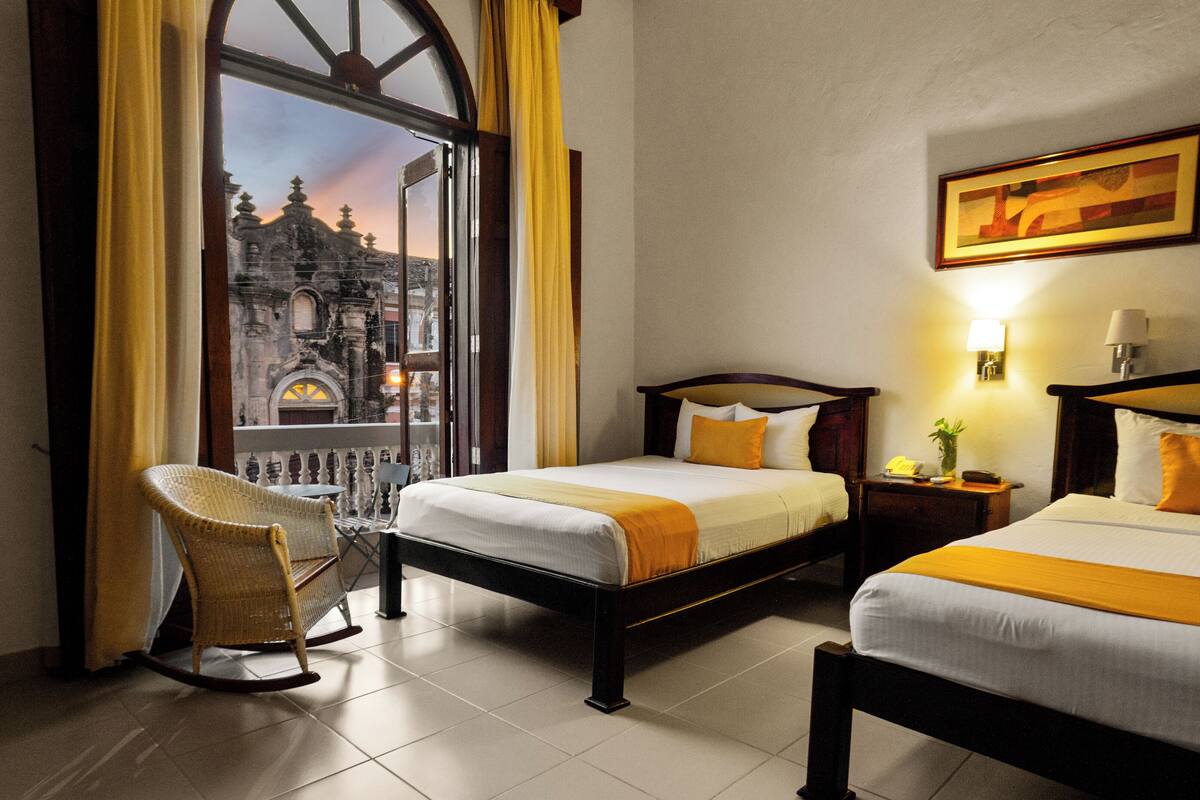 Quarto de hotel com duas camas, uma cadeira de balanço, vista para um edifício histórico e um quadro na parede.