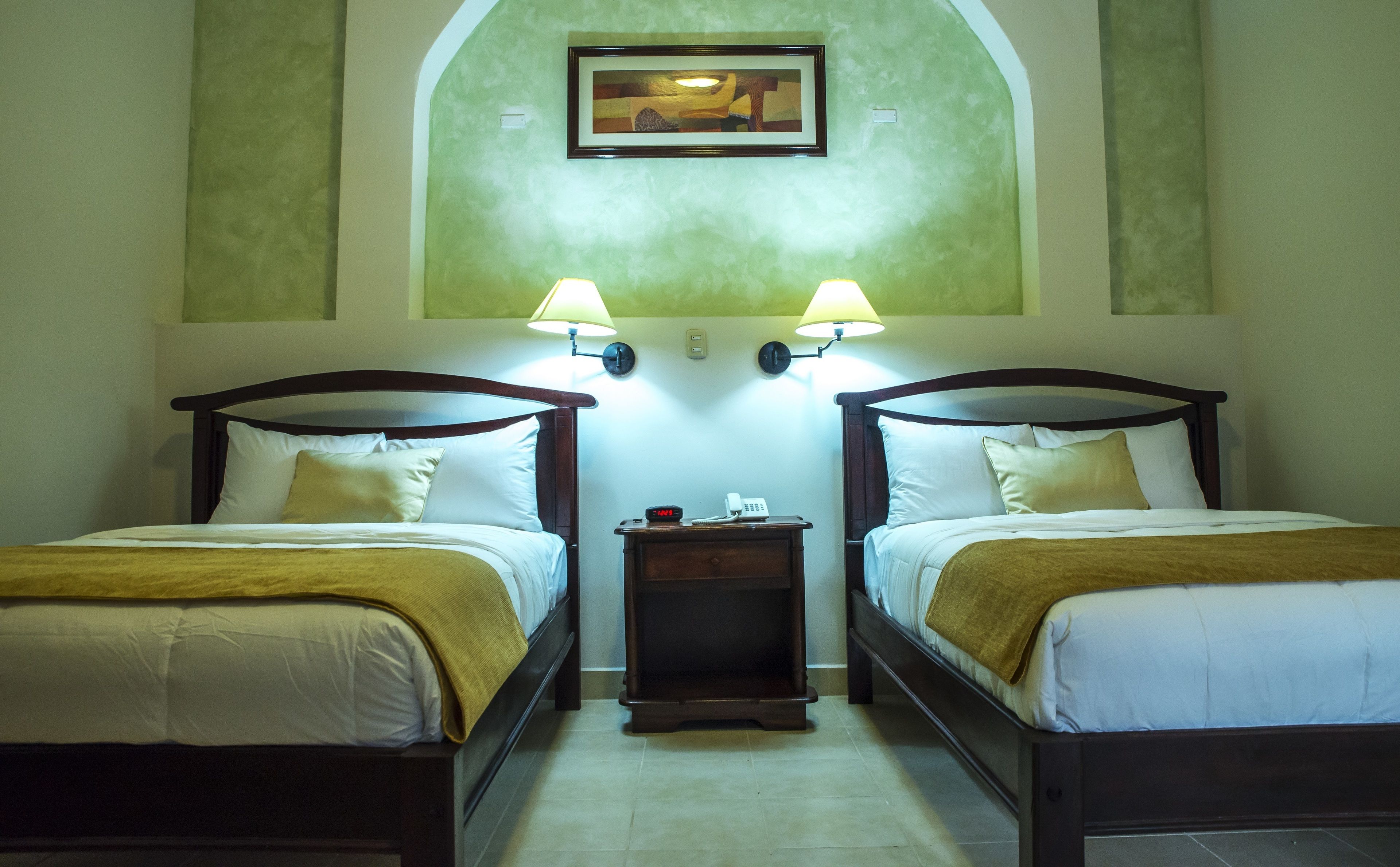 A hotel room with two single beds, each with a wooden headboard, white bedding, and a brown throw blanket. There are two bedside lamps, a small wooden nightstand with a telephone and a clock, and a framed picture above the beds.