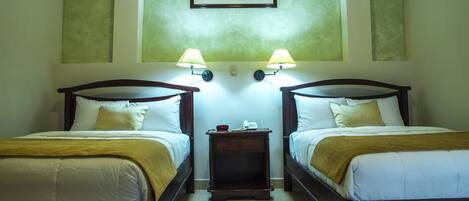 Habitación de hotel con dos camas individuales, cada una con cabecero de madera, ropa de cama blanca y una manta marrón. Hay dos lámparas de noche, una mesita de noche de madera con un teléfono y un reloj, y un cuadro enmarcado sobre las camas.