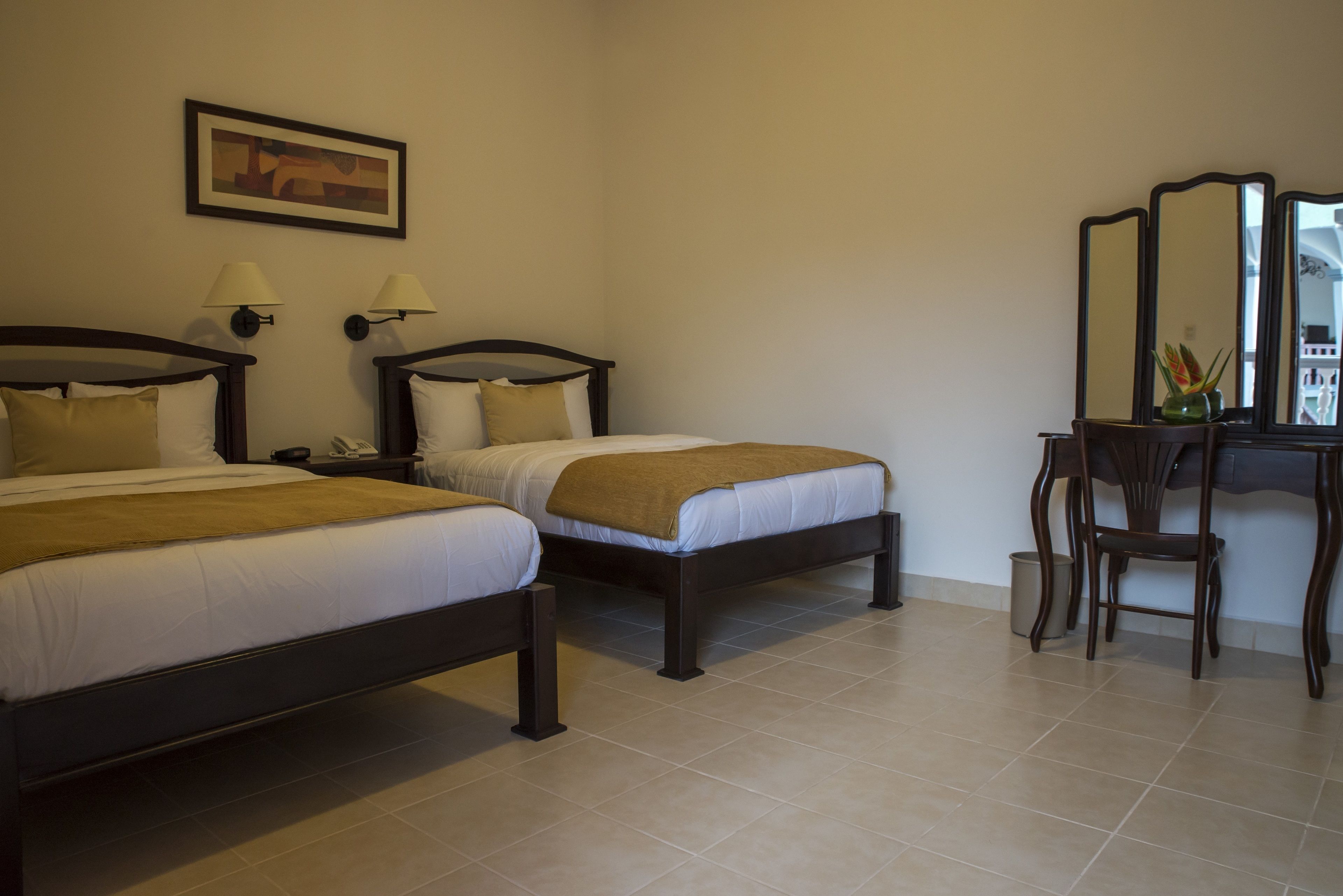 Quarto de hotel com duas camas, uma escrivaninha, uma cadeira, um espelho e um quadro na parede.