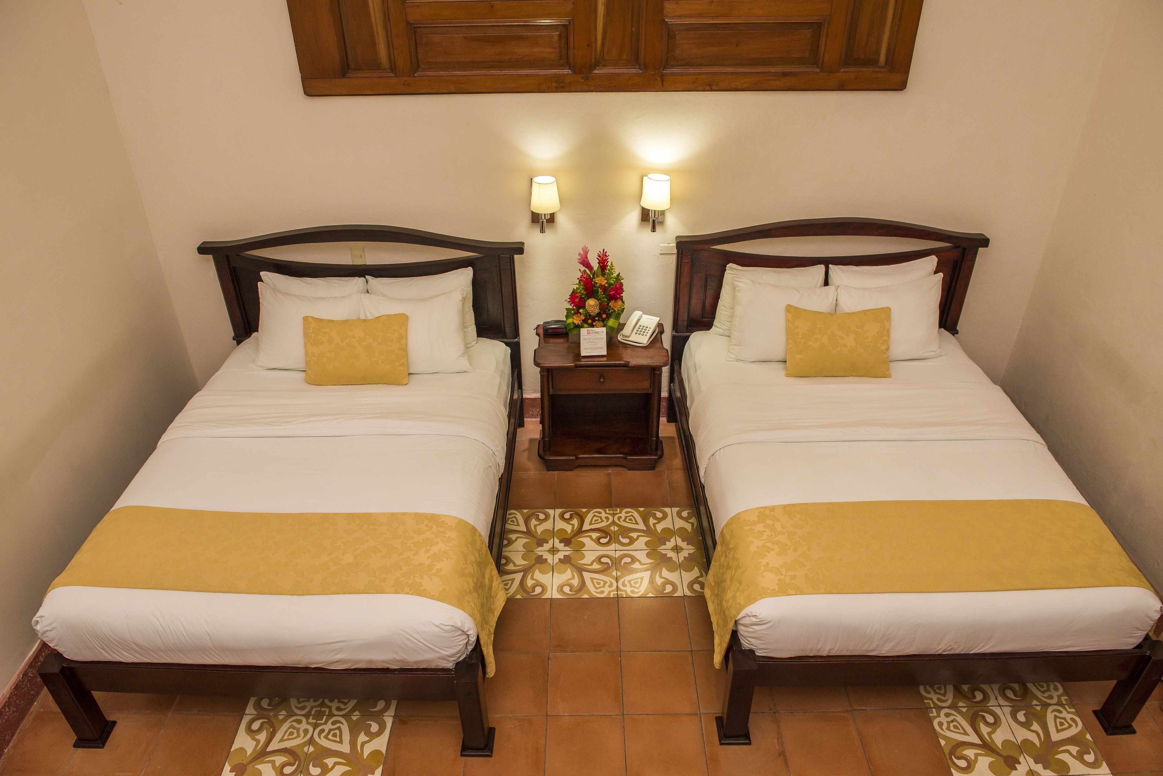 A hotel room with two beds, each with a wooden headboard, white bedding, and a yellow throw. There are two bedside lamps and a small table with a telephone and a floral arrangement.