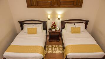 A hotel room with two beds, each with a wooden headboard, white bedding, and a yellow throw. There are two bedside lamps and a small table with a telephone and a floral arrangement.