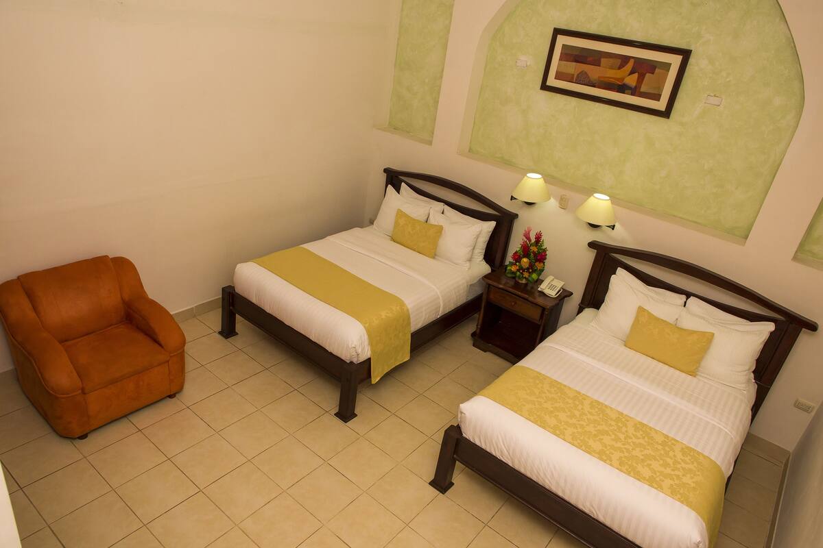 Quarto de hotel com duas camas, uma cadeira e uma mesinha de cabeceira com luminárias.