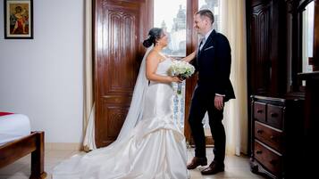 Una pareja de novios en una habitación con cama, armario de madera y una puerta que da al exterior.
