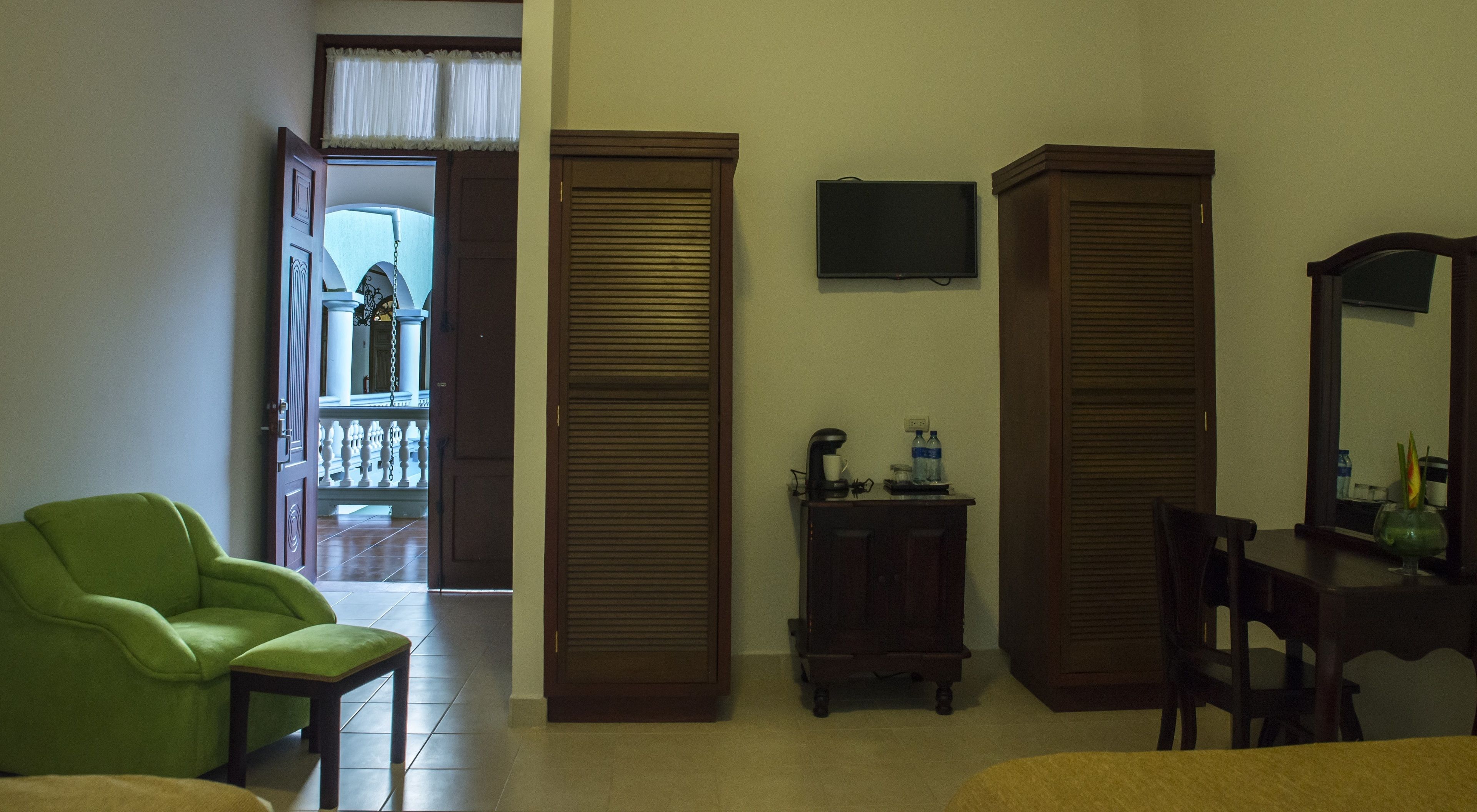 A hotel room with a green armchair, a wooden cabinet with a coffee maker, and a small table with a mirror.