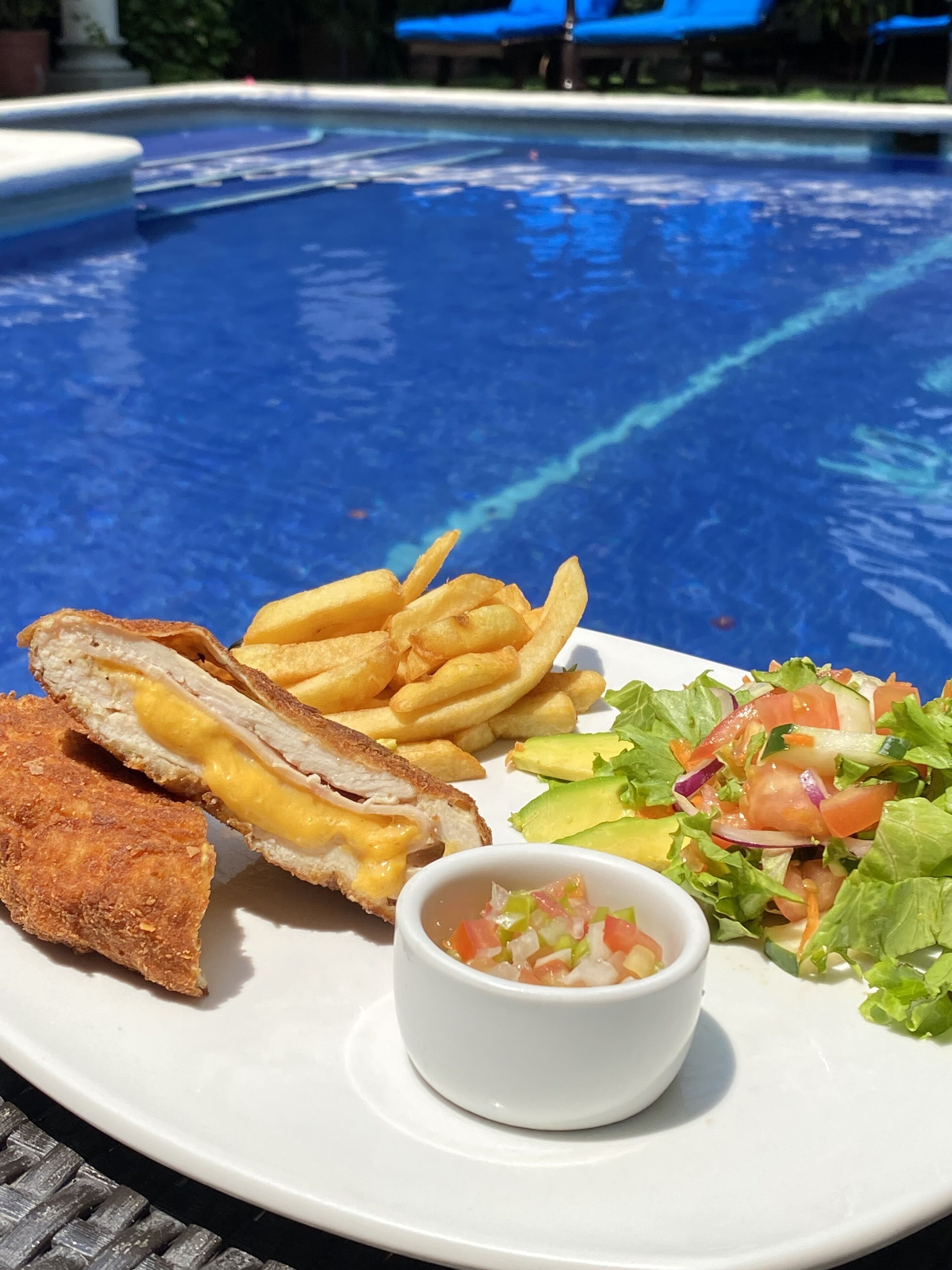 Ein Teller mit Essen, darunter ein Sandwich, Pommes, Salat und eine kleine Schale Salsa, vor einer Poolkulisse.