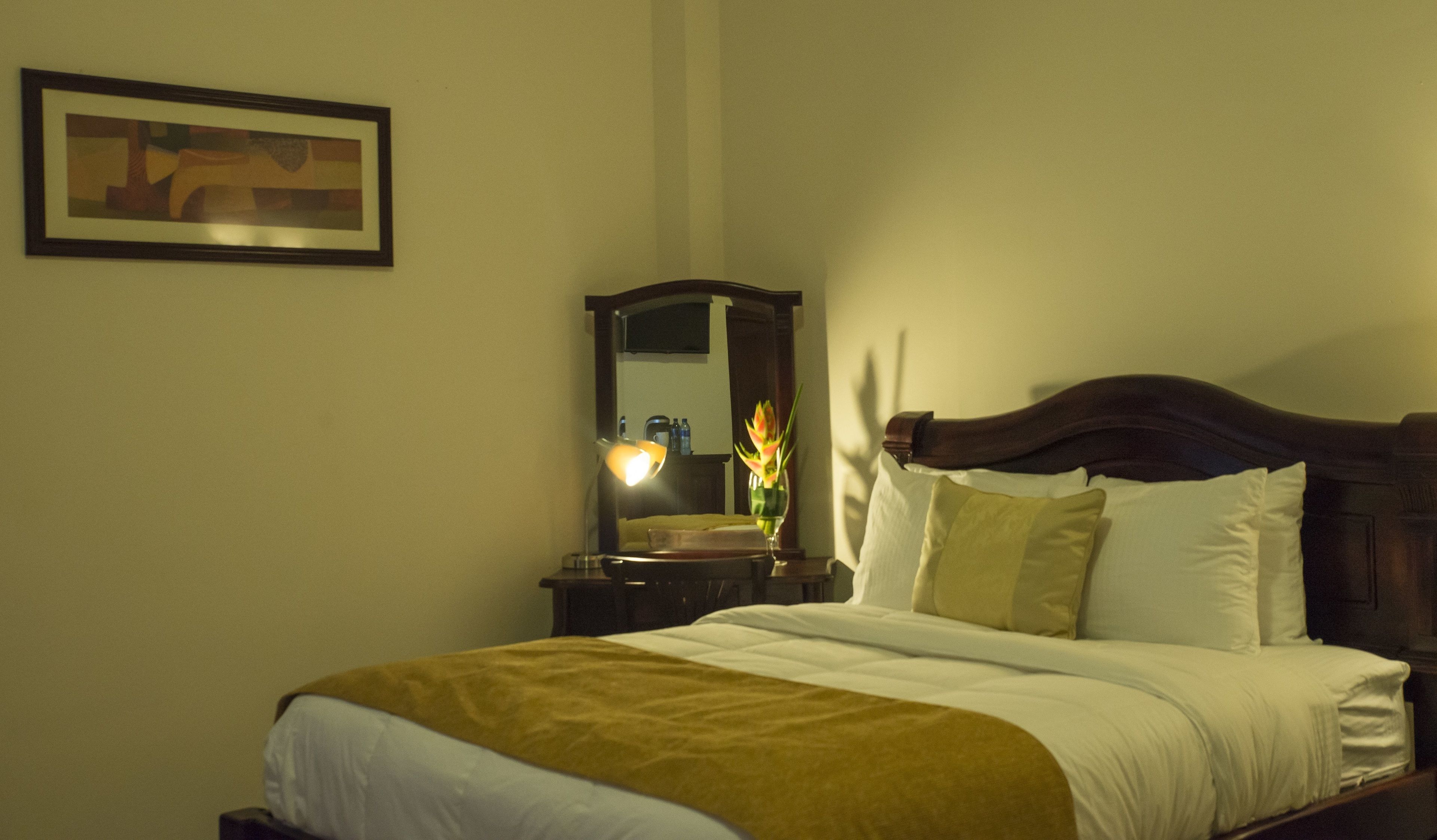A neatly made bed with white and yellow bedding, a wooden headboard, and a bedside table with a lamp and a vase of flowers.