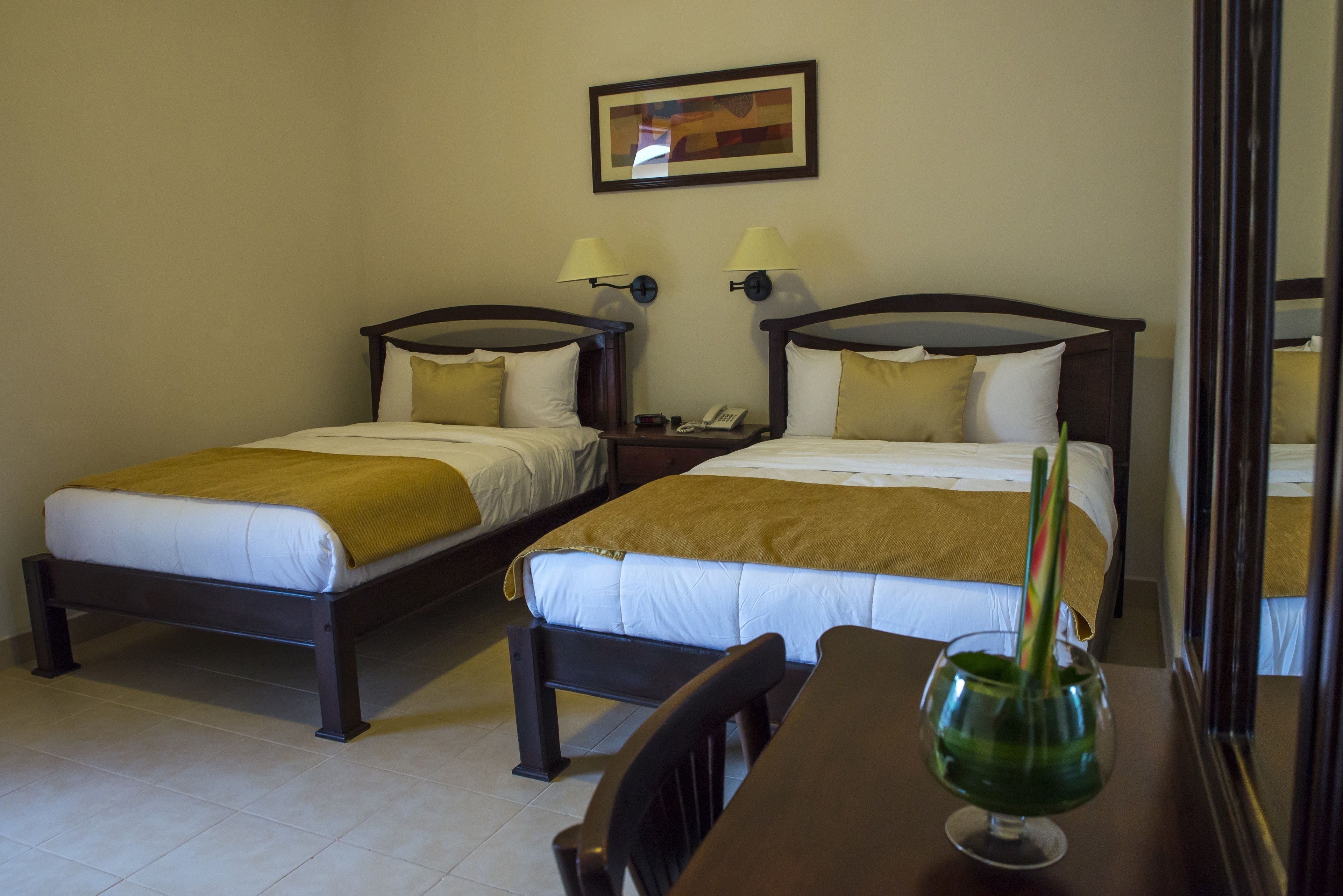 A hotel room with two single beds, each with a wooden frame and white bedding. There are two bedside tables with lamps, a small table with a glass and a green plant, and a framed picture on the wall.