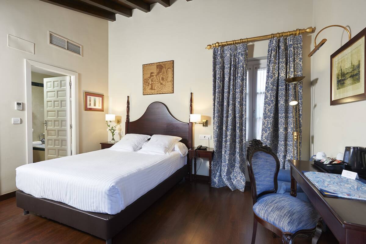 Una cama bien hecha con sábanas blancas, cabecera de madera, mesita de noche con lámpara, una silla con cojín estampado y un escritorio con una computadora portátil y papeles.