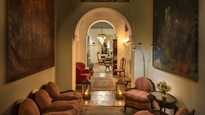 Ein Flur mit einem roten Sessel, einer gemusterten Couch und einem kleinen Tisch mit einer Vase. Zwei Laternen stehen auf dem Boden und ein Chandelier ist im Hintergrund zu sehen.