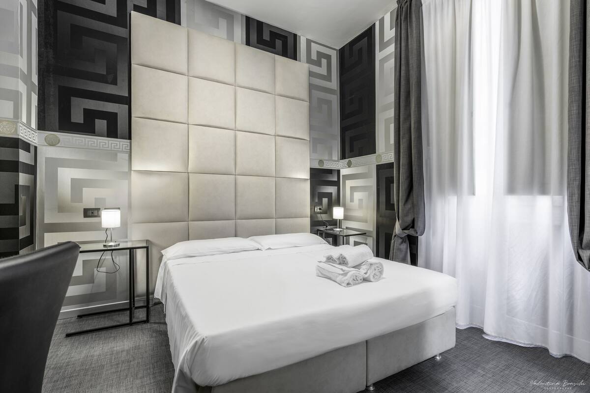 A modern hotel room with a large bed, a bedside table with a lamp, a chair, and a mirror.