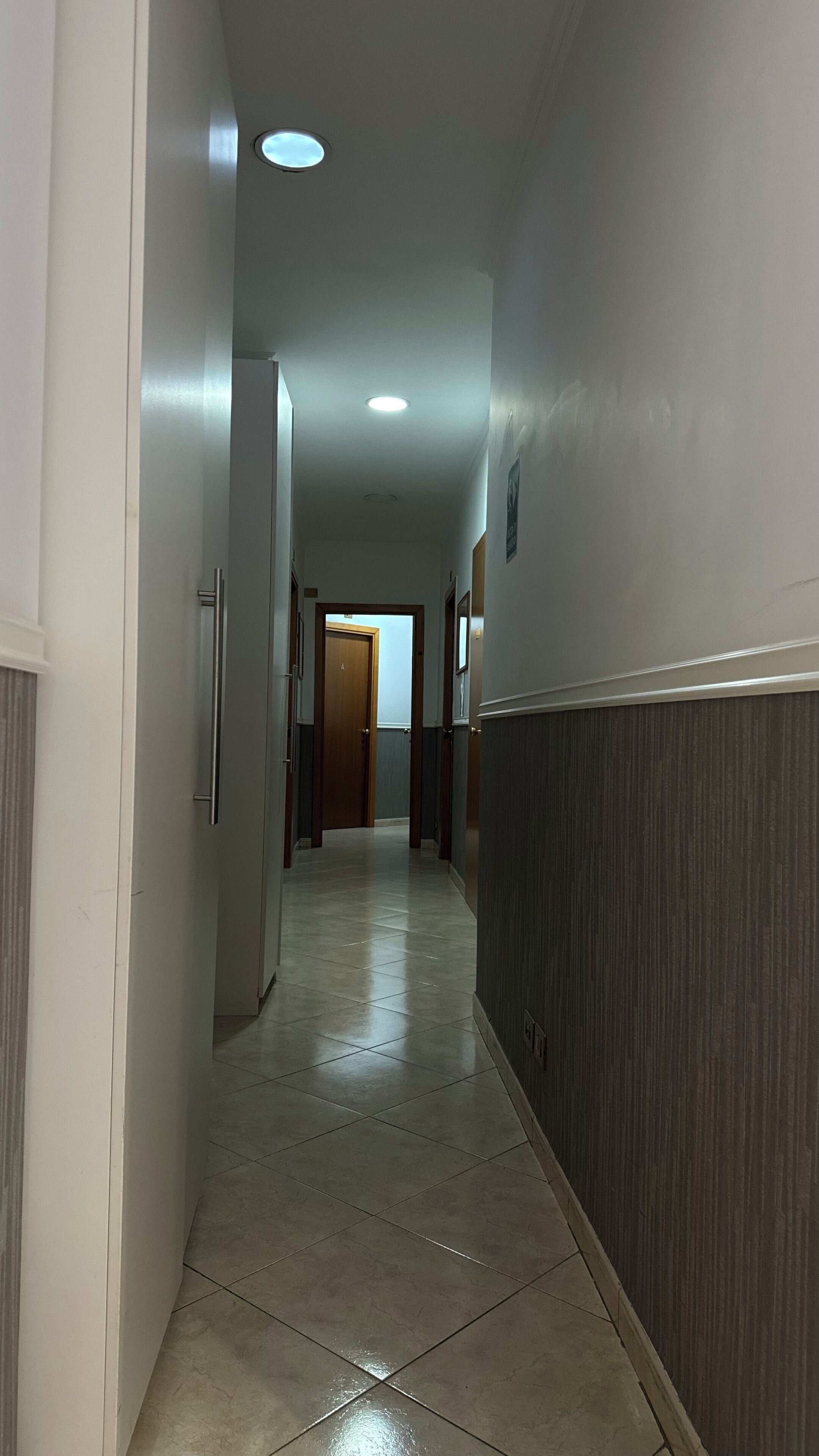 Um corredor com piso de azulejos, uma porta de madeira no final e um luminário no teto.