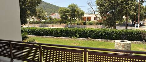 A balcony with a metal railing overlooking a well-maintained garden, a stone wall, and a street with buildings and cars.