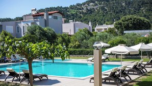A swimming pool with lounge chairs and umbrellas, surrounded by greenery and buildings. - Santa Helena Hotel Ialysos (Rhodes)