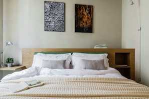 A neatly made bed with white bedding, a bedside table with a lamp, and two framed artworks on the wall.
