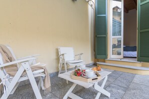 A balcony with two white chairs, a small table with a tray of food and drinks, and a window with green shutters.