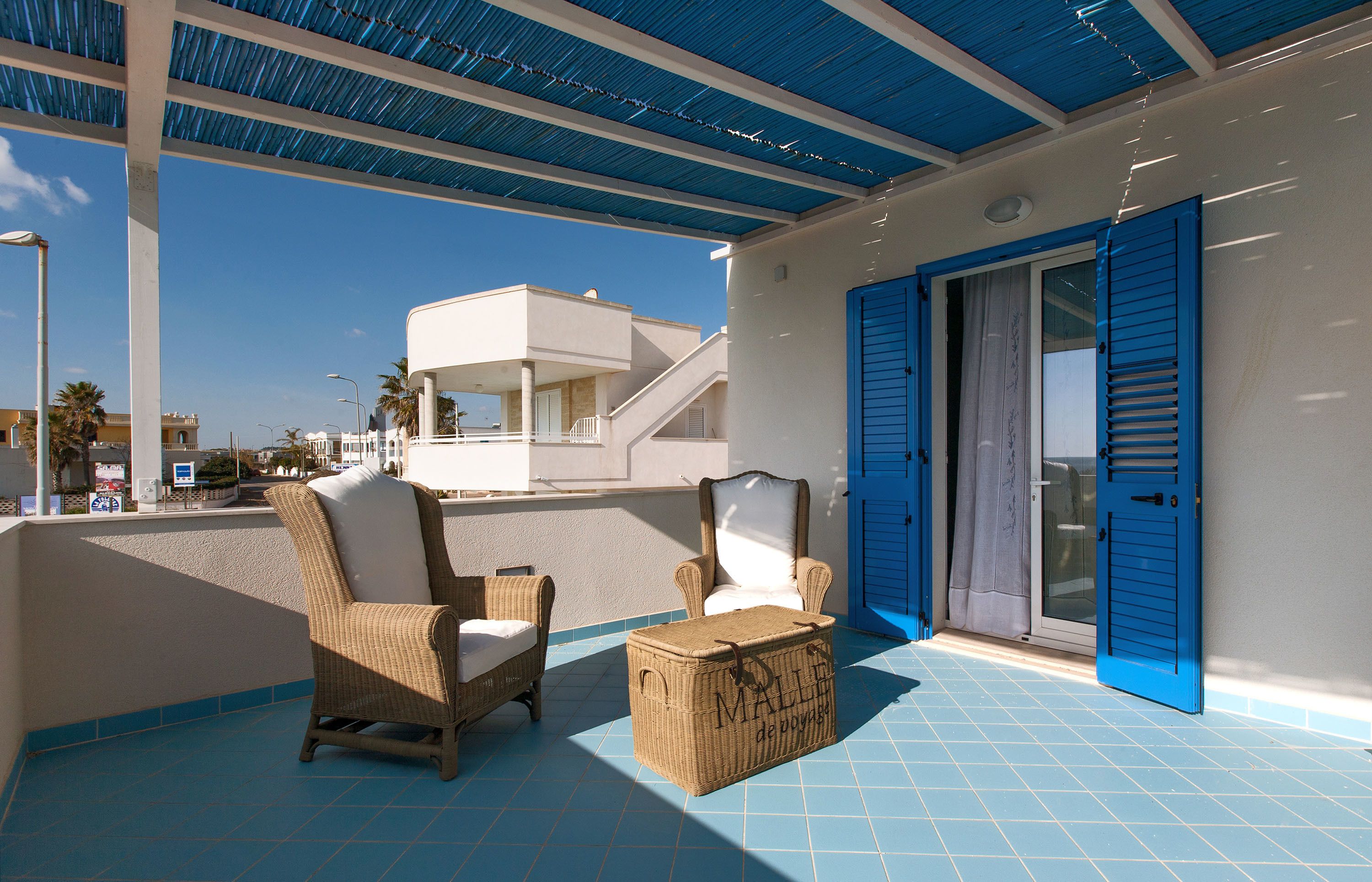 Un espace extérieur abrité, aménagé avec des meubles et un coffre en osier, une porte bleue et une vue sur un bâtiment blanc en arrière-plan.