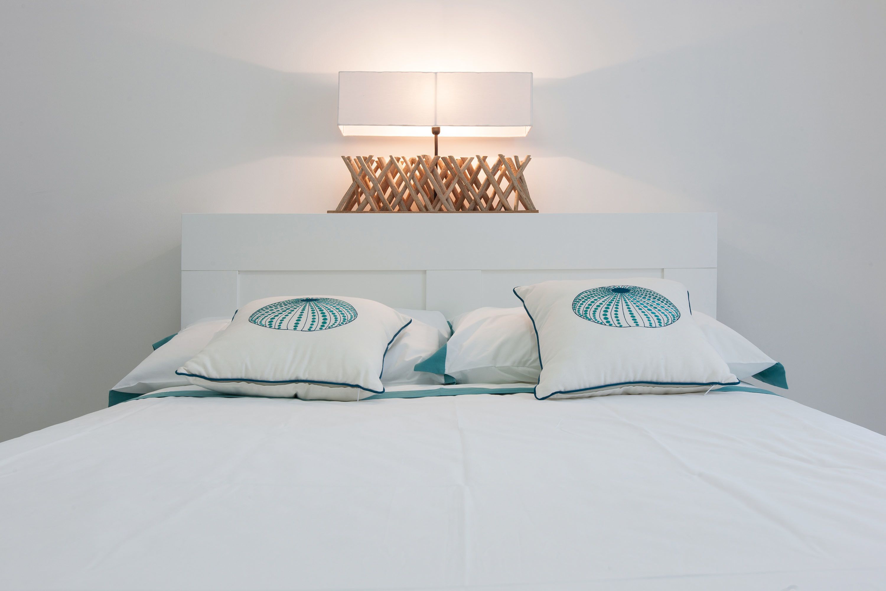 A neatly made bed with white bedding and teal pillows, a white headboard, and a wall-mounted lamp with a wooden lattice design.