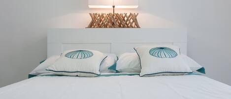 A neatly made bed with white bedding and teal pillows, a white headboard, and a wall-mounted lamp with a wooden lattice design.