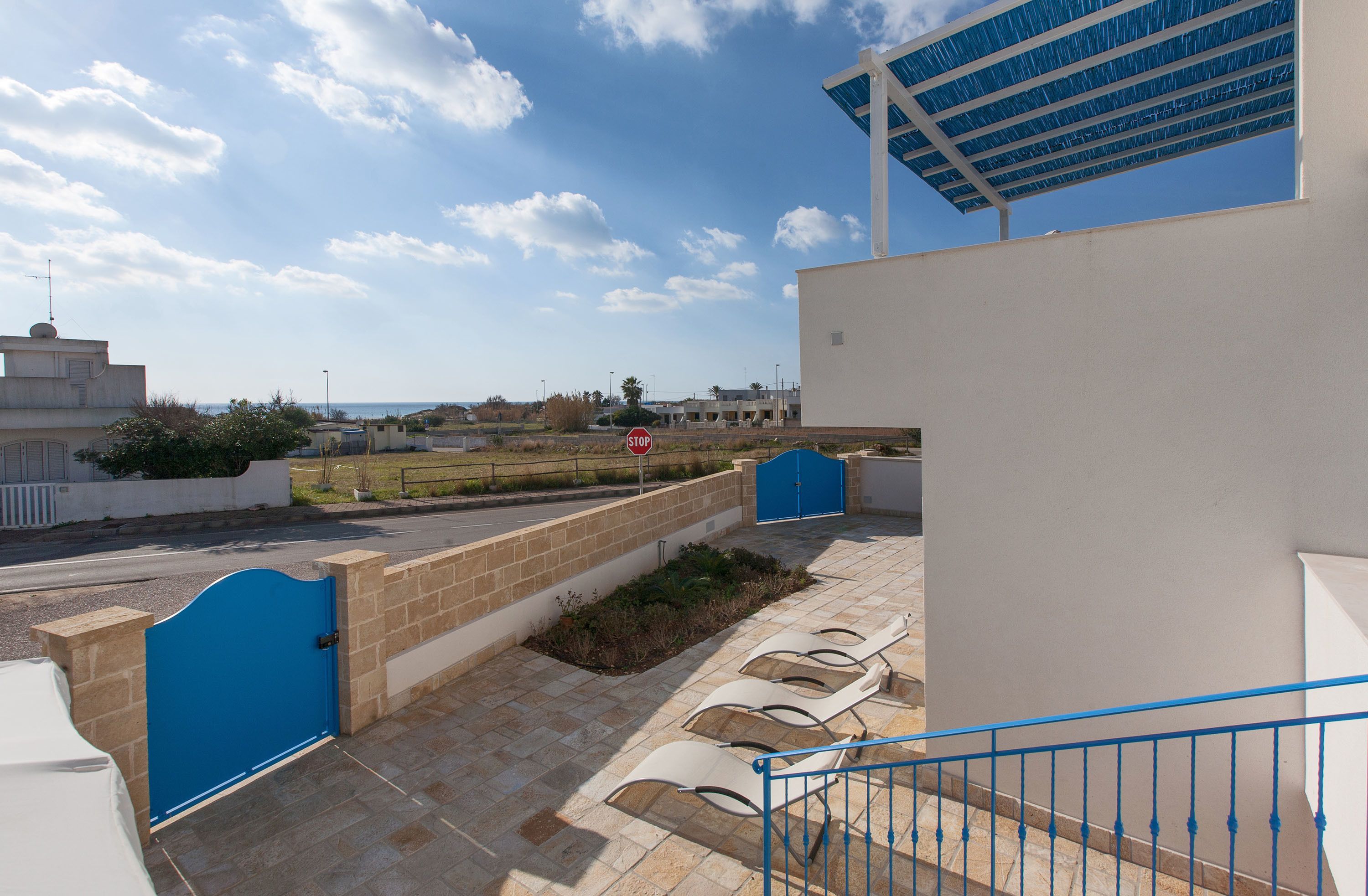 Una entrada con puerta azul y reja, un espacio de estar al aire libre cubierto, y vista al mar y edificios a lo lejos.