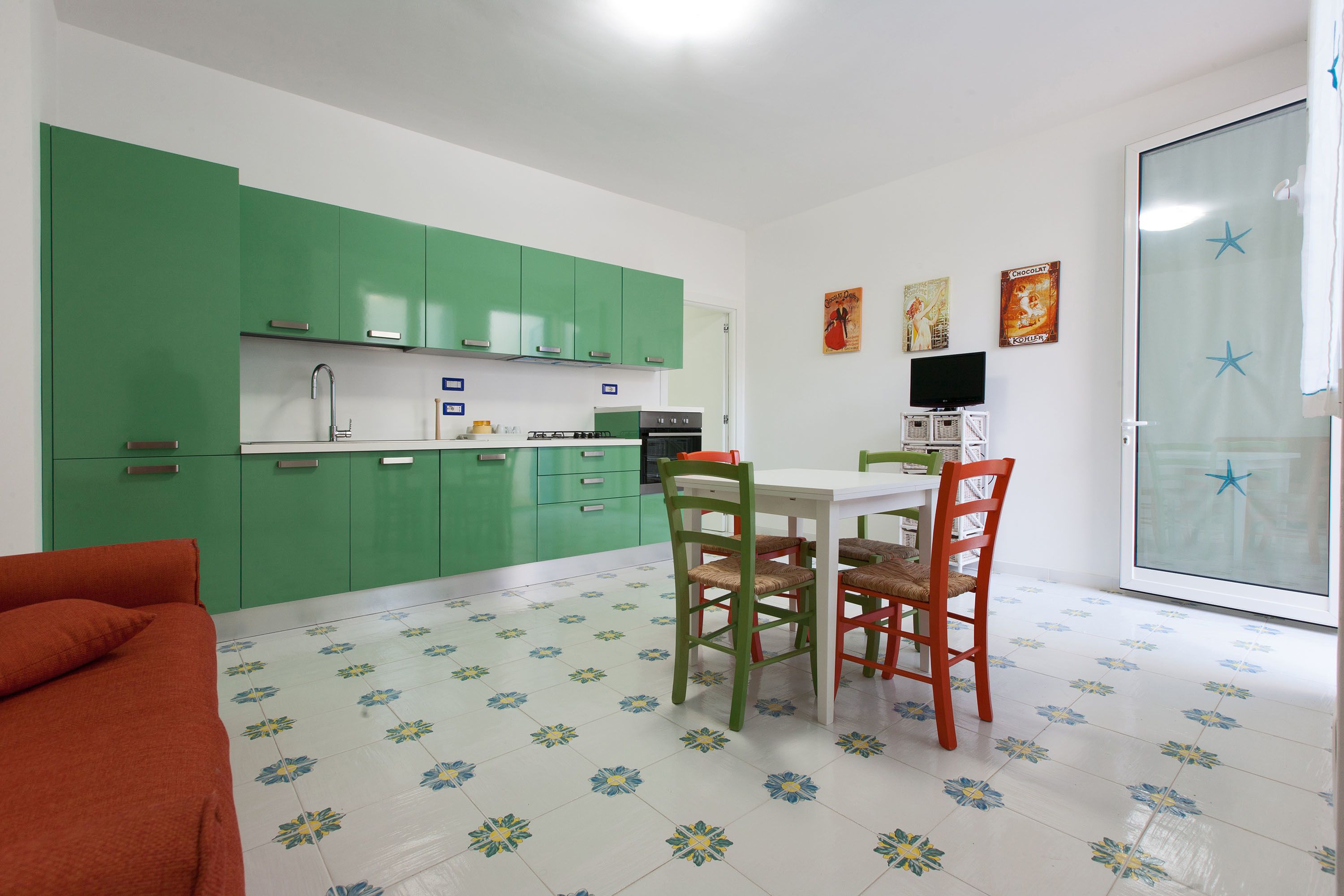 A compact kitchen with green cabinets, a dining table with two chairs, and a television on a stand.