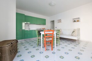 Egy kis méretű konyha, zöld szekrényekkel és egy fehér étkezőasztallal, melyhez narancssárga székek tartoznak.