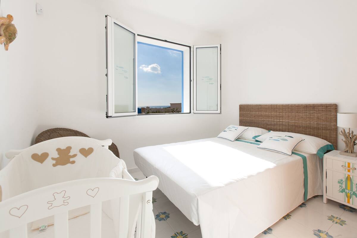 Ein Schlafzimmer mit einem Bett, einem Babybett, einem Nachttisch und einem Fenster mit freier Aussicht.