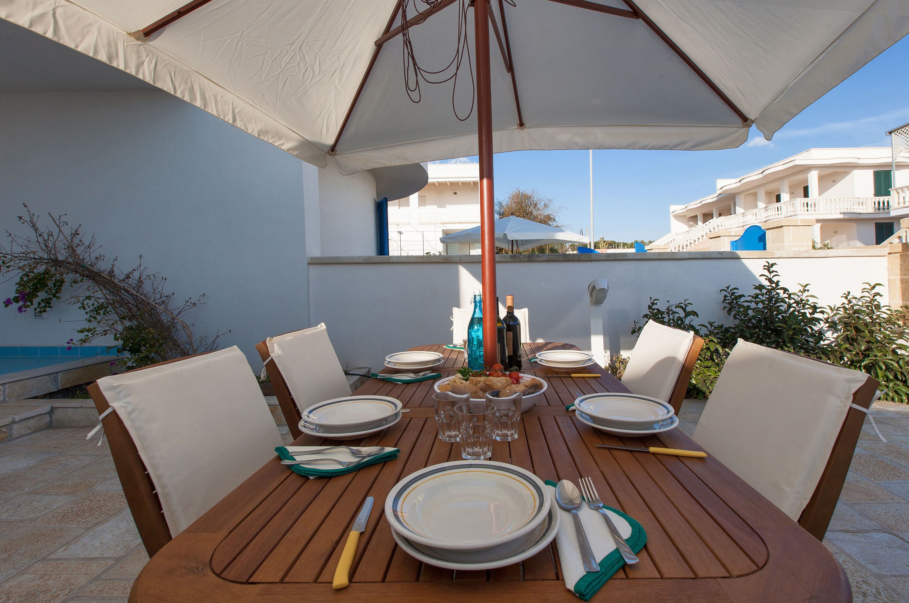 Uma mesa de jantar de madeira posta com pratos, talheres e copos, sob um guarda-sol grande, com uma piscina e edifícios ao fundo.