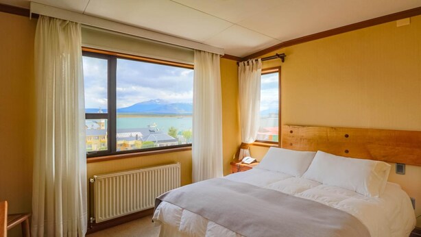 A hotel room with a large bed, a wooden headboard, a view of a lake and mountains, and a window with curtains. - Hotel Saltos del Paine (Natales)