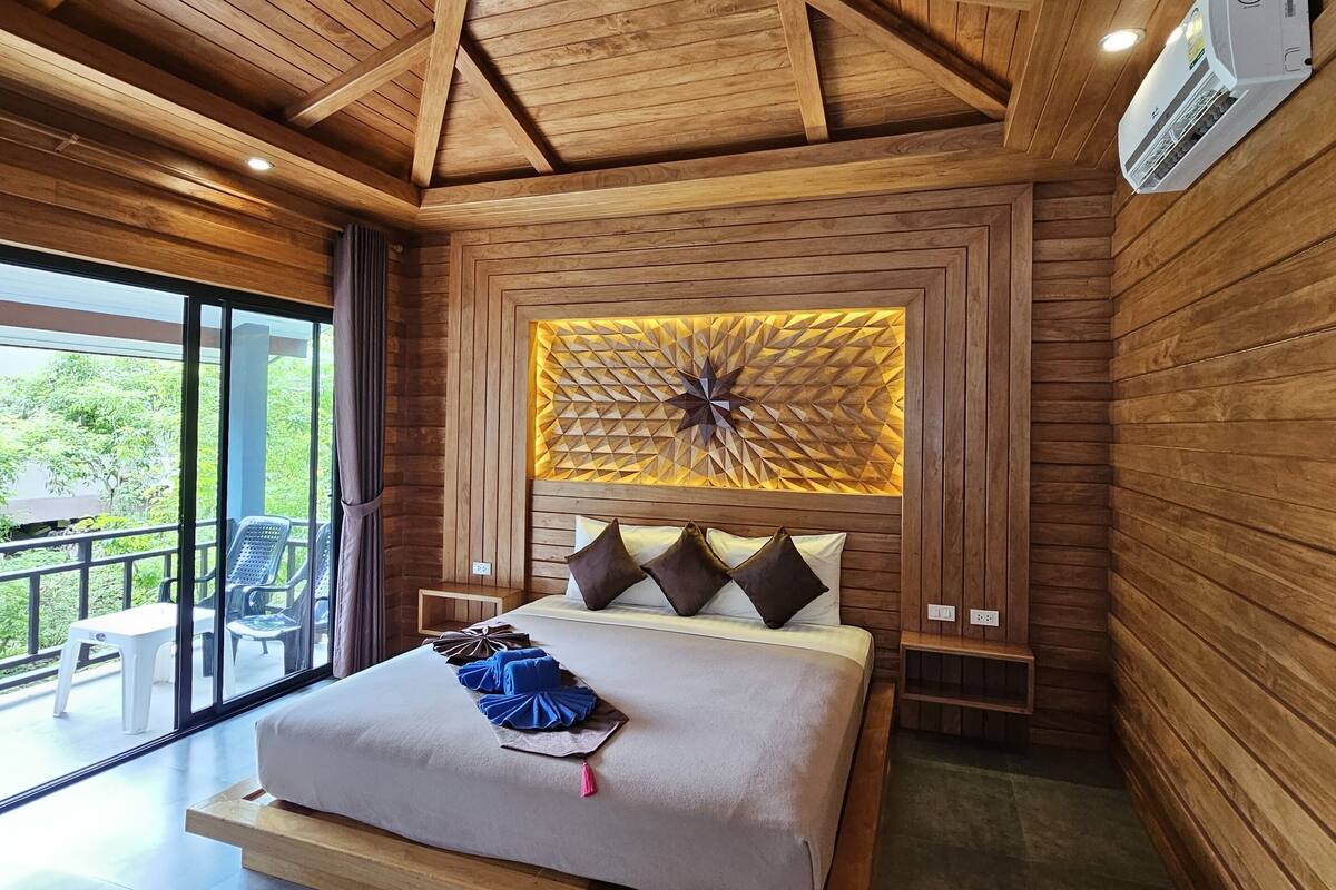 A spacious bedroom with a wooden ceiling, a large bed with a headboard, and a view of the outdoors through a sliding glass door.
