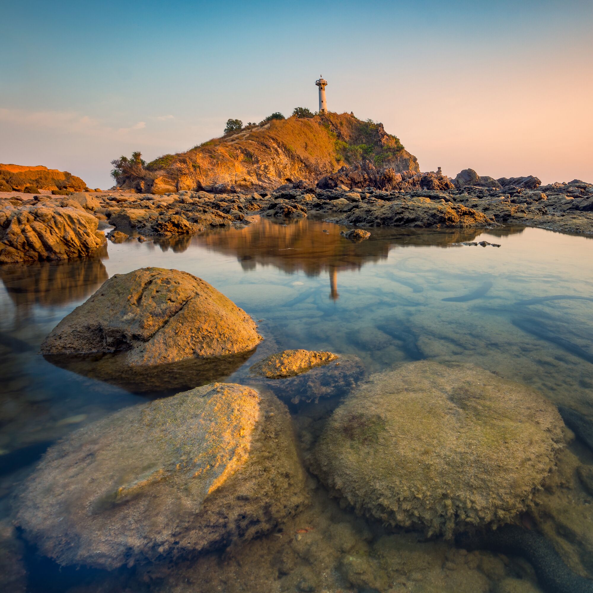 Uma paisagem costeira com um farol numa colina rochosa, água cristalina refletindo as rochas, e um céu calmo.