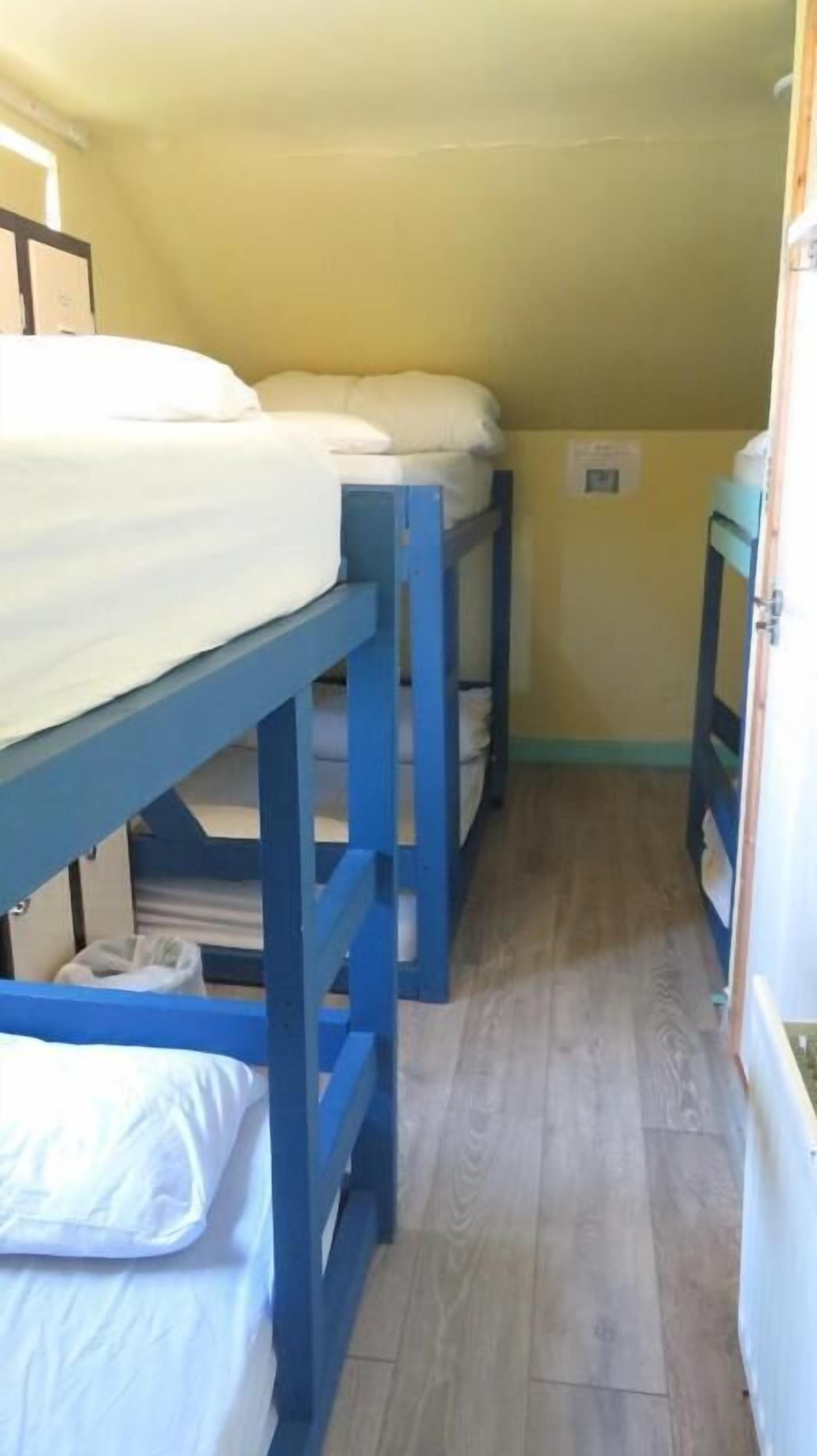 Un couloir étroit, avec des lits superposés alignés le long des murs, un plancher en bois et une porte sur le côté droit.