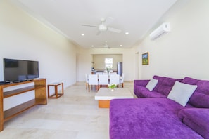 Ein modernes Wohnzimmer mit einem violetten Sofa, einem Fernseher auf einem Holzgestell und einem Essbereich im Hintergrund.