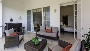 A spacious living area with a sofa, chairs, a coffee table, and a dining table. There is a sliding glass door leading to a bedroom with a bed and a nightstand.