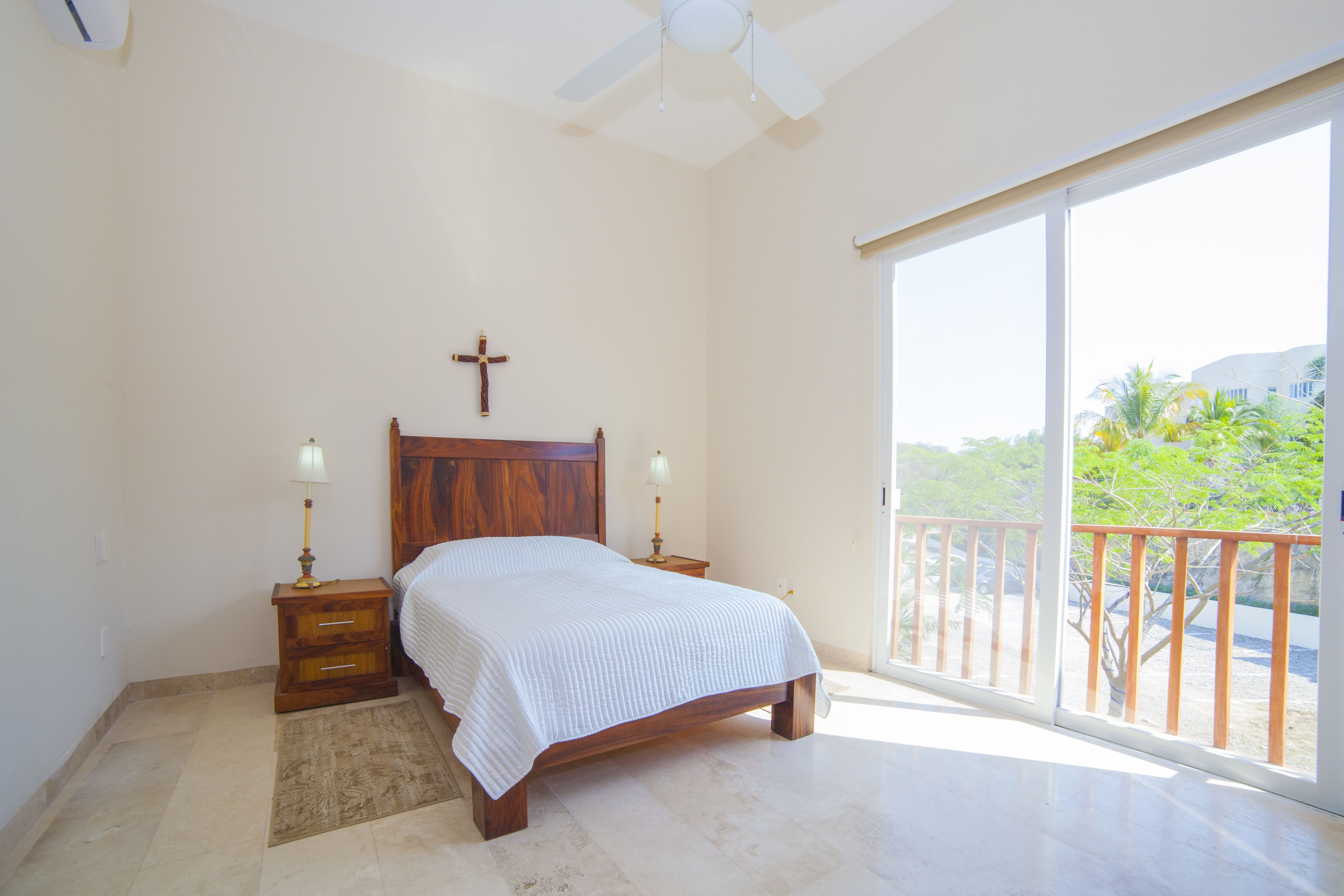 A bedroom with a wooden bed, two bedside tables with lamps, a cross on the wall, and a balcony with a view of greenery.