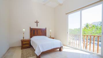 A bedroom with a wooden bed, two bedside tables with lamps, a cross on the wall, and a balcony with a view of greenery.