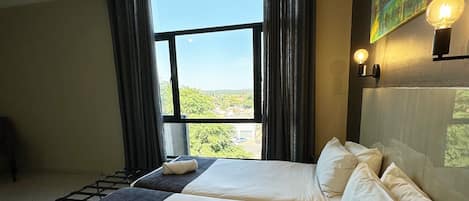 A hotel room with two beds, a large window with curtains, a painting on the wall, and a wall-mounted light fixture.