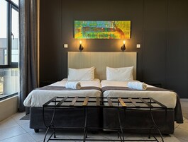 Una cama doble con cabecera, dos mesitas de noche con lámparas, un cuadro abstracto grande sobre la cama y vistas al exterior a través de una ventana con cortinas.