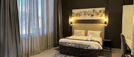 A hotel room with a bed, a bedside table, a chair, a large window with curtains, and a wall-mounted artwork.