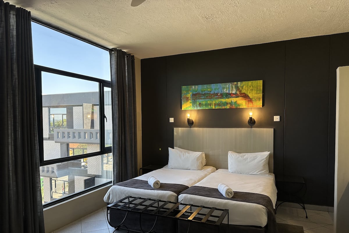 Een hotelkamer met twee bedden, een groot raam met gordijnen, een schilderij aan de muur en een klein tafeltje met dienblad.