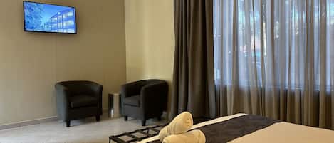 A hotel room with a bed, two chairs, a TV, and a window with curtains.