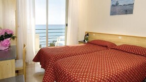 A bedroom with a bed, a desk, a chair, a nightstand, a lamp, and a window with a view of the sea. - Hotel Savoia (Ospedaletti)