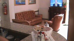 A living room with a brown leather sofa, a television displaying a news program, and a small table with a basket of bread. - Hotel Savoia (Ospedaletti)
