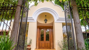 Una puerta doble de madera con un panel de vidrio, flanqueada por dos portones metálicos, que conduce a un edificio con un arco blanco y un aplique colgante sobre la puerta.