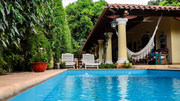 Una piscina con sillas de descanso blancas y una hamaca, rodeada de vegetación exuberante y una estructura de techo de tejas.