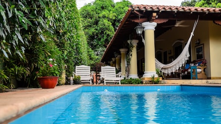 A swimming pool with white lounge chairs and a hammock, surrounded by lush greenery and a tiled roof structure.